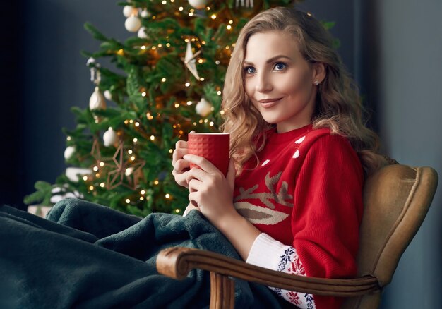 Portrait of gorgeous blonde woman in red sweater chilling under a plaid with cup of coffee in the Christmas decorated interior.