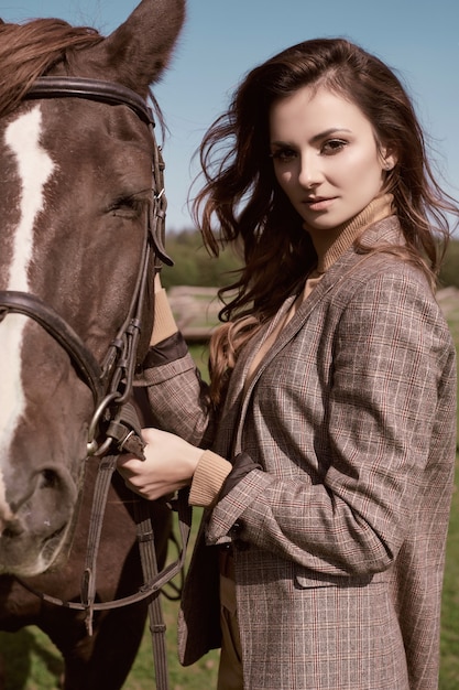 Portrait of a gorgeous brunette woman in an elegant checkered brown jacket posing with a horse on country landscape