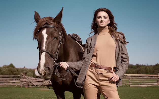 Portrait of a gorgeous brunette woman in an elegant checkered brown jacket posing with a horse on country landscape