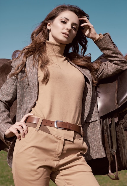 Portrait of a gorgeous brunette woman in an elegant checkered brown jacket posing with a horse on country landscape