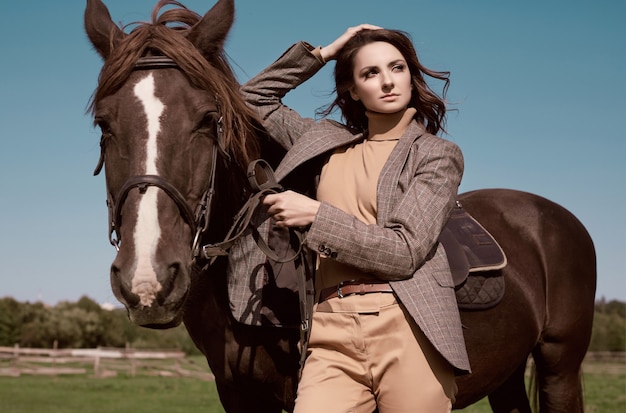 Portrait of a gorgeous brunette woman in an elegant checkered brown jacket posing with a horse on country landscape