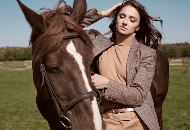 Portrait of a gorgeous brunette woman in an elegant checkered brown jacket posing with a horse on country landscape