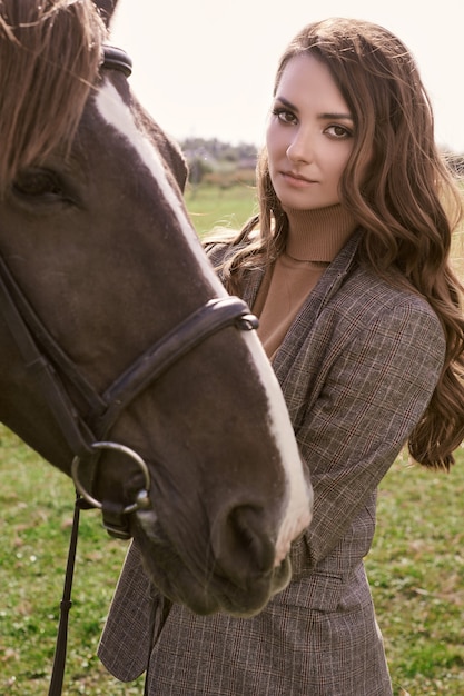 Portrait of a gorgeous brunette woman in an elegant checkered brown jacket posing with a horse on country landscape