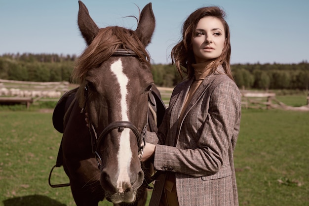 Portrait of a gorgeous brunette woman in an elegant checkered brown jacket posing with a horse on country landscape