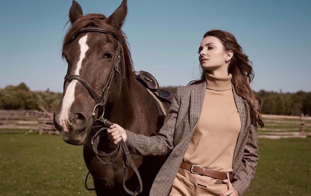 Portrait of a gorgeous brunette woman in an elegant checkered brown jacket posing with a horse on country landscape