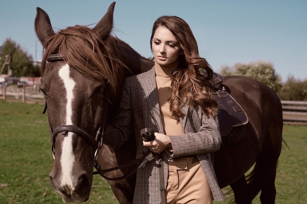 Portrait of a gorgeous brunette woman in an elegant checkered brown jacket posing with a horse on country landscape