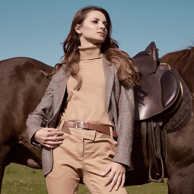 Portrait of a gorgeous brunette woman in an elegant checkered brown jacket posing with a horse on country landscape