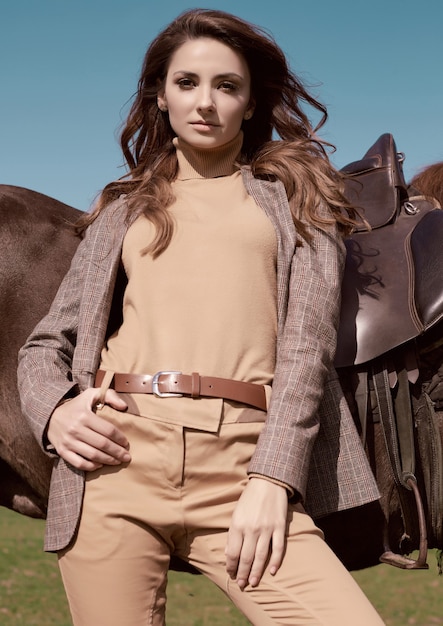 Portrait of a gorgeous brunette woman in an elegant checkered brown jacket posing with a horse on country landscape