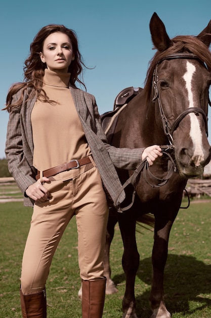 Portrait of a gorgeous brunette woman in an elegant checkered brown jacket posing with a horse on country landscape