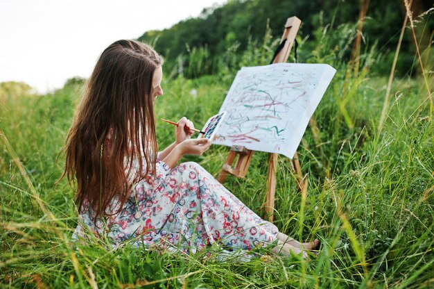 Portrait of a gorgeous happy young woman in beautiful dress sitting on the grass and painting on paper with watercolors.