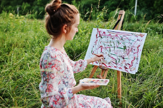 Portrait of a gorgeous happy young woman in beautiful dress sitting on the grass and painting on paper with watercolors