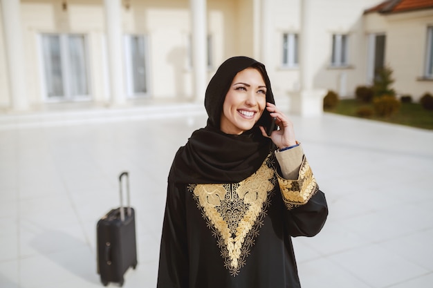 Portrait of gorgeous muslim woman in traditional wear using smart phone for calling taxi while standing in front of her house. In background luggage.