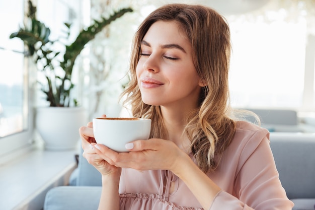 Portrait of gorgeous smiling lady smelling and drinking cappuccino from cup, while resting in restaurant in morning