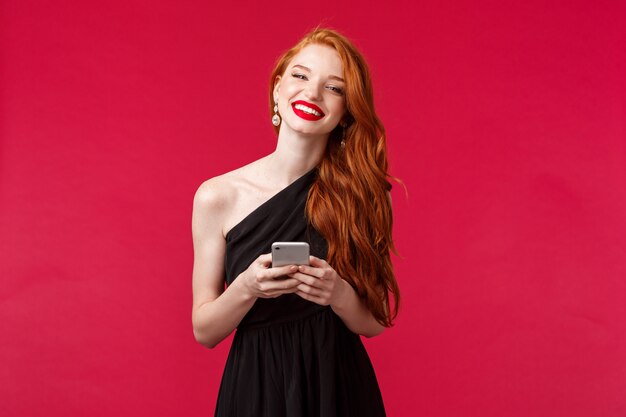 Portrait of gorgeous smiling woman with ginger hair in elegant black prom dress, holding smartphone, messaging, using application, want take picture, laughing and looking , on a red wall