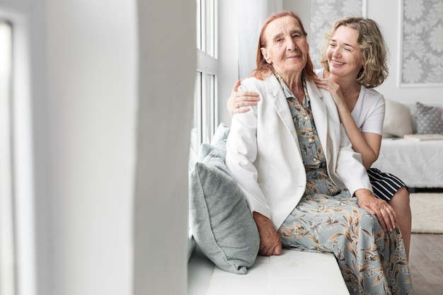 Portrait of granddaughter and grandmother sitting on window sill at home