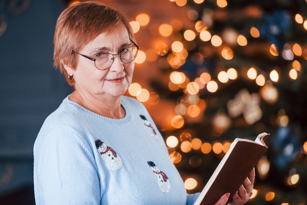 Portrait of grandmother that standing in New year decorated room with book.