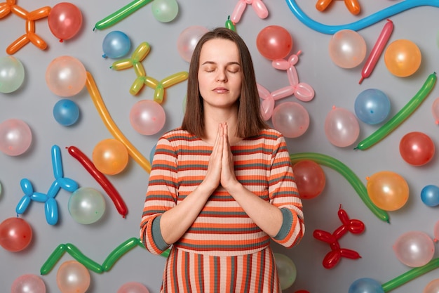 Portrait of grateful calm woman with brown hair wearing striped dress posing over gray background with inflated balloons relaxing with yoga praying before festive holiday dinner