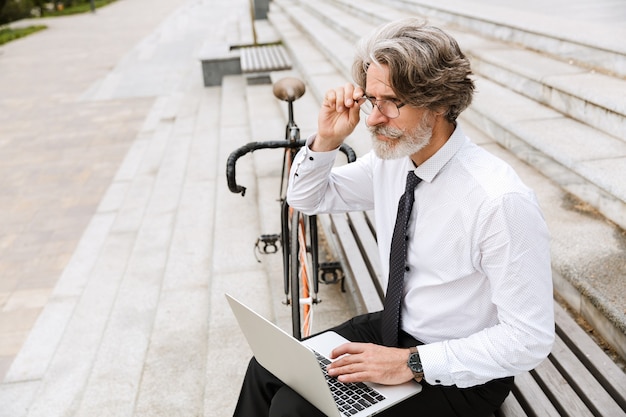 Portrait of gray-haired adult businessman in eyeglasses typing on laptop while sitting on bench with bicycle outdoors