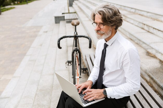 Portrait of gray-haired mature businessman in eyeglasses typing on laptop while sitting on bench with bicycle outdoors