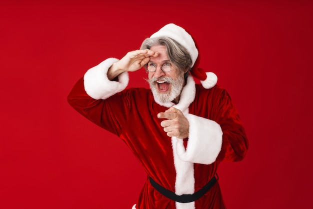 Portrait of gray-haired Santa Claus old man in costume looking and pointing finger at copyspace isolated on red