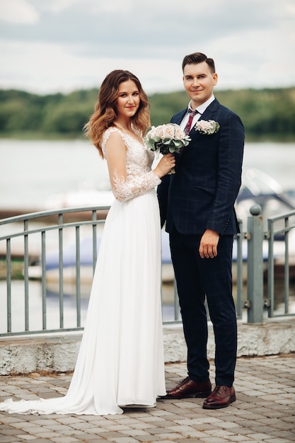 Portrait of groom in black suit with beautiful bride in white dress in the park stands near the lake