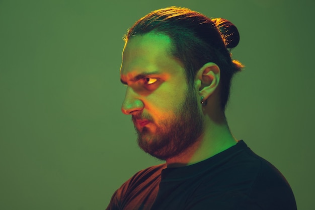 Portrait of a guy with colorful neon light on green  wall. Male model with calm and serious mood. Facial expression, millenials lifestyle and look like. Future, technologies.