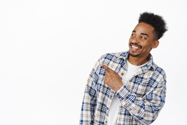 Portrait of handsome african american guy smiling happy, pointing and looking left at sale promo banner, showing advertisement, standing in casual clothes on white.