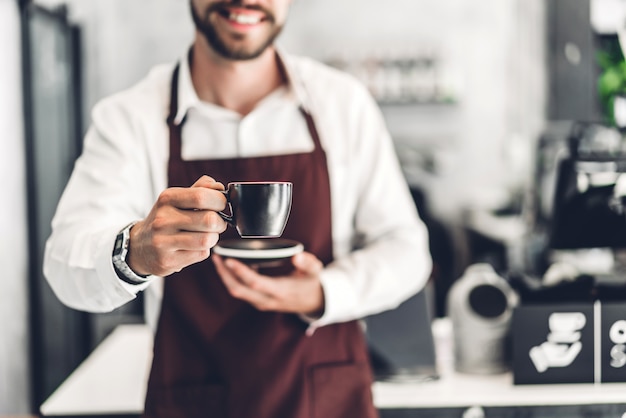 Portrait of handsome bearded barista man small smiling and holding cup of coffee in the cafe