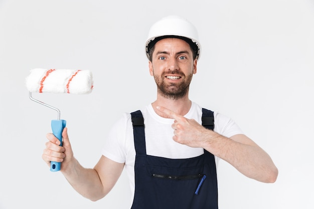 Portrait of a handsome bearded builder man wearing overalls standing isolated over white wall, holding paint brush