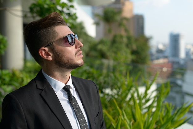 Portrait of handsome bearded Hispanic businessman with nature in the city outdoors