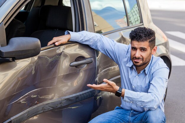 Portrait of handsome bearded man wearing jeans and shirt sitting near auto and showing with hands dents and scratches on the door of his automobile, damage car after road accident, Outdoor shot.