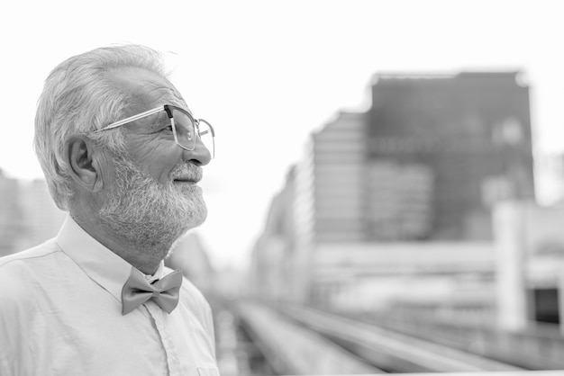 Portrait of handsome bearded senior tourist man wearing stylish clothes while exploring the city of Bangkok in black and white