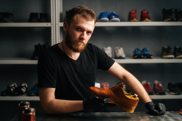 Portrait of handsome bearded shoemaker examining light brown leather shoes during restoration working