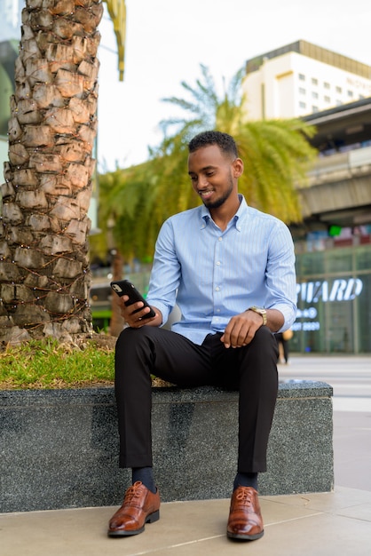 Portrait of handsome black African businessman outdoors in city during summer using mobile phone while smiling vertical shot