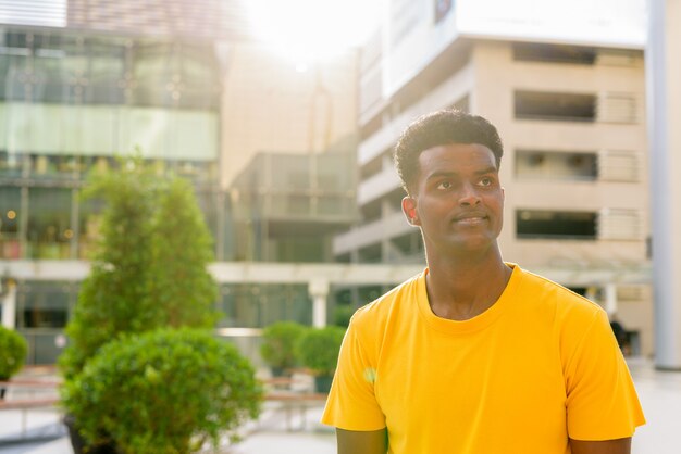 Portrait of handsome black African man wearing yellow t-shirt outdoors in city during summer backlit shot with lens flare while thinking