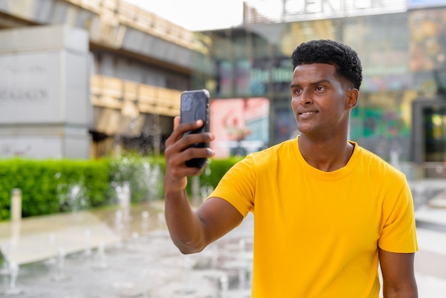 Portrait of handsome black African man wearing yellow t-shirt outdoors in city during summer next to fountain while using mobile phone