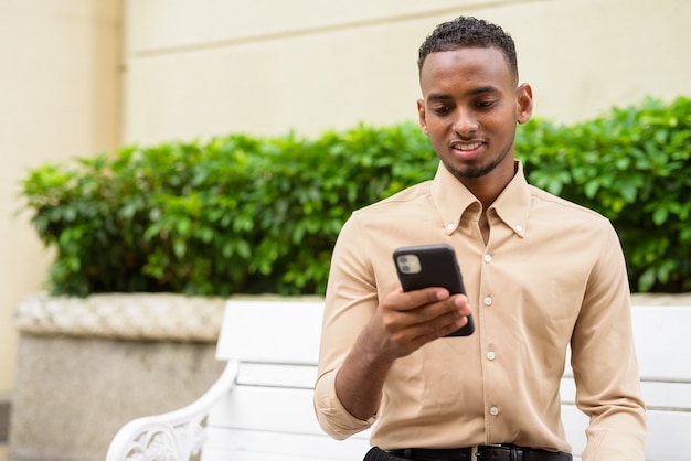 Portrait of handsome black young African businessman wearing casual clothes outdoors in city and using mobile phone