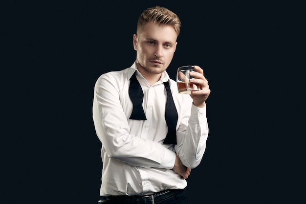 Portrait of handsome blond man in elegant tuxedo and bow tie posing with glass of whiskey on black  wall