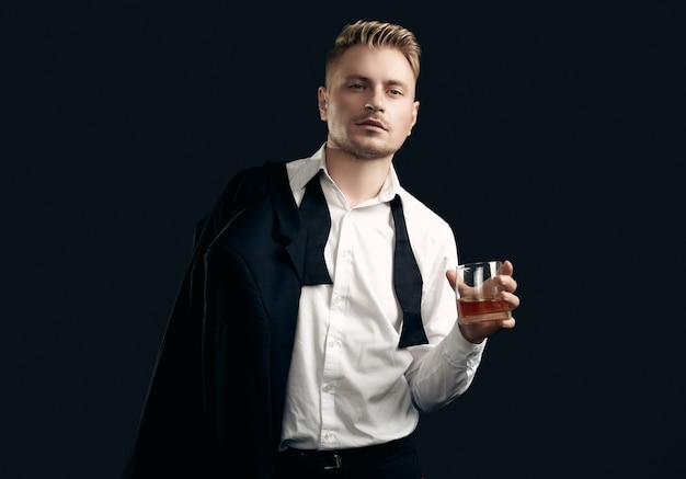 Portrait of handsome blond man in elegant tuxedo and bow tie posing with glass of whiskey on black  wall