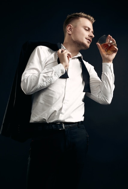 Portrait of handsome blond man in elegant tuxedo and bow tie posing with glass of whiskey on black  wall