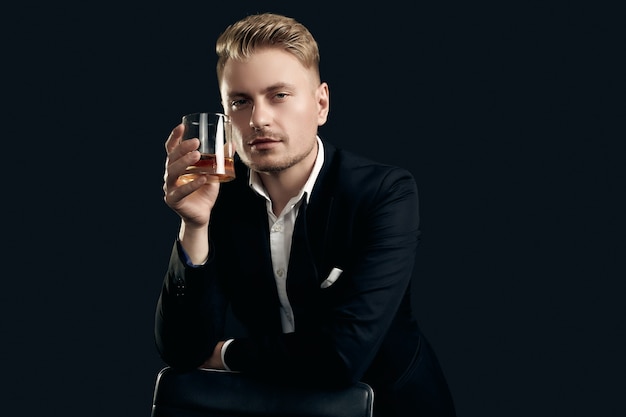 Portrait of handsome blond man in elegant tuxedo and bow tie posing with glass of whiskey on black  wall