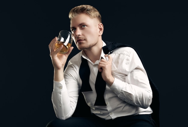 Portrait of handsome blond man in elegant tuxedo and bow tie posing with glass of whiskey on black  wall