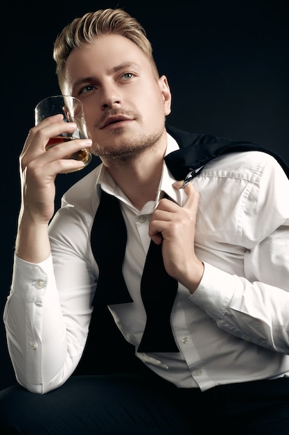 Portrait of handsome blond man in elegant tuxedo and bow tie posing with glass of whiskey on black  wall