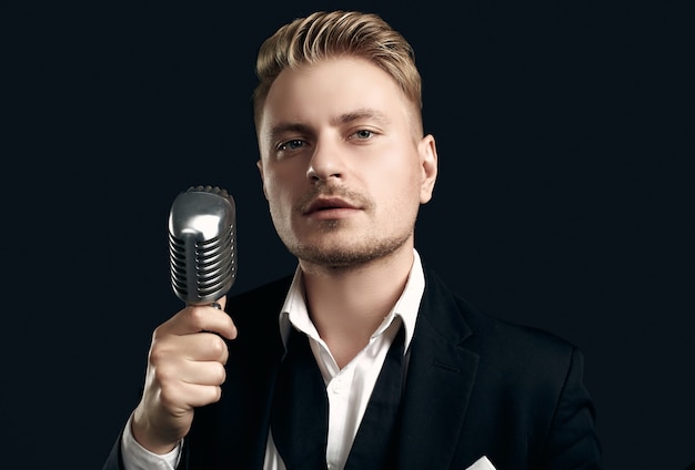 Portrait of handsome blond man singer in elegant tuxedo and bow tie posing with vintage microphone on black  wall