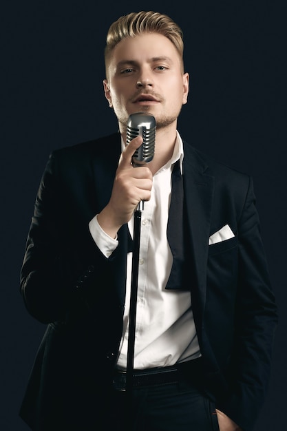 Portrait of handsome blond man singer in elegant tuxedo and bow tie posing with vintage microphone on black  wall