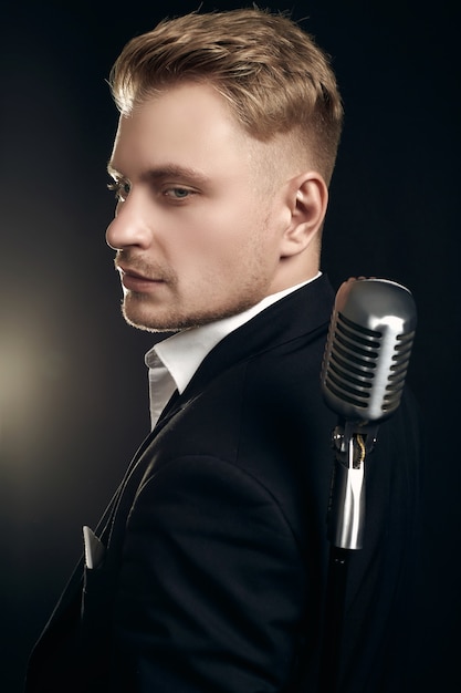 Portrait of handsome blond man singer in elegant tuxedo and bow tie posing with vintage microphone on black  wall