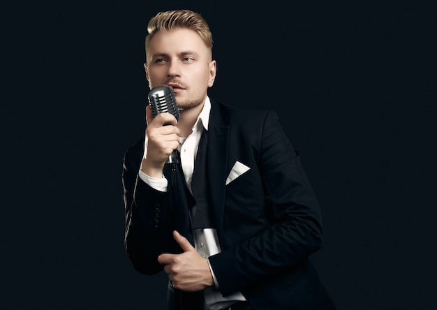 Portrait of handsome blond man singer in elegant tuxedo and bow tie posing with vintage microphone on black  wall