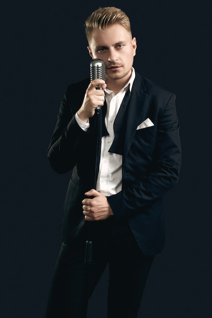 Portrait of handsome blond man singer in elegant tuxedo and bow tie posing with vintage microphone on black  wall