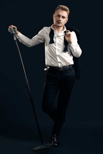 Portrait of handsome blond man singer in elegant tuxedo and bow tie posing with vintage microphone on black  wall