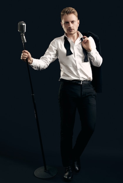 Portrait of handsome blond man singer in elegant tuxedo and bow tie posing with vintage microphone on black  wall